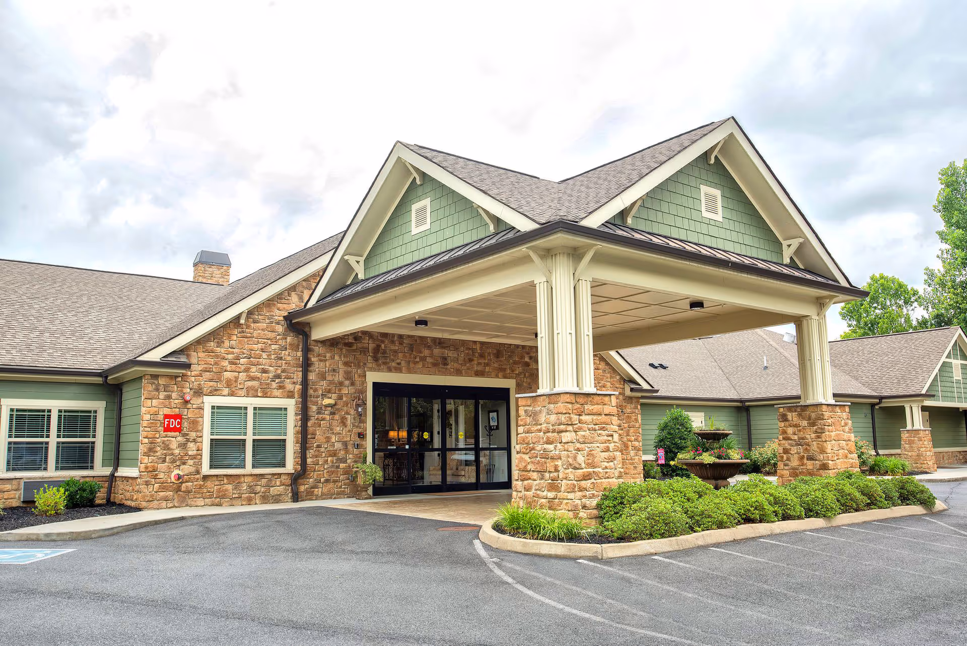 Exterior view of Dominion Senior Living of Sevierville featuring a covered entrance with stone pillars, green siding, and a paved driveway with landscaped bushes and a fountain.