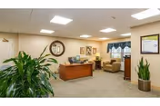 A well-lit office reception area with a wooden desk, computer, and office chair. There are two comfortable armchairs near a window with blue valance curtains. The walls are decorated with framed pictures and a round mirror. Two large potted plants are placed on either side of the room.