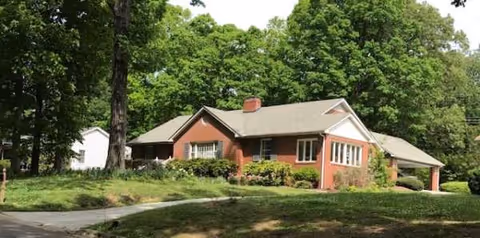 Brick, single-story building with a gabled roof amid lush trees.