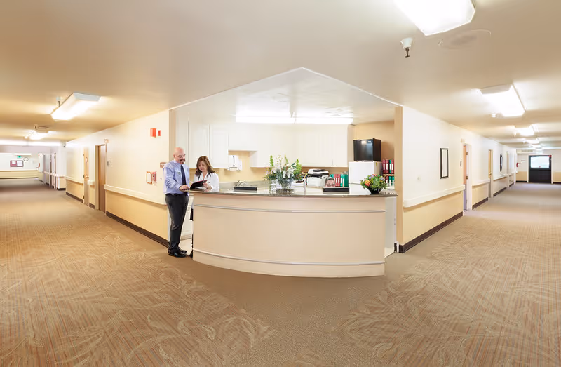 A wide hallway in a senior living facility with beige walls and carpeted floors. In the center is a reception desk with two staff members standing behind it, one man and one woman, looking at a clipboard. The hallway extends on both sides with doors and handrails along the walls. The area is well-lit with ceiling lights.