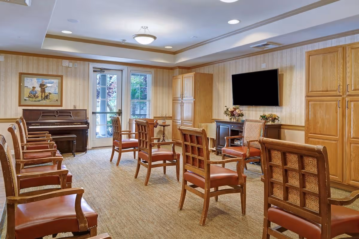 Well-lit common sitting room with arranged wooden chairs, a piano, a wall-mounted TV, and wooden cabinets.