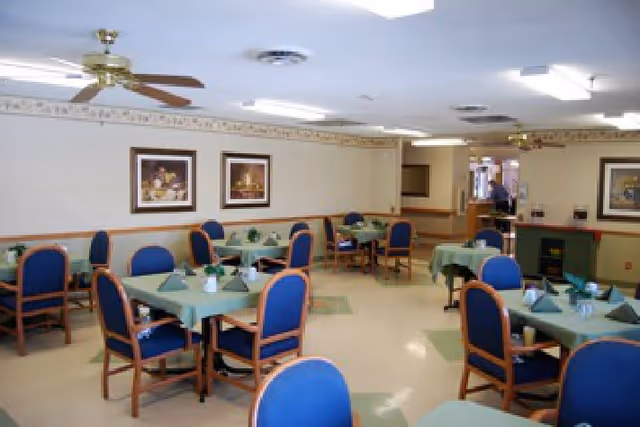 A dining room with several tables covered in green tablecloths, each surrounded by blue cushioned chairs with wooden frames. The tables are set with folded green napkins and small potted plants. The room has ceiling fans, fluorescent lighting, framed artwork on the walls, and a beige and green color scheme.