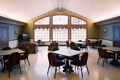 A spacious common area with several tables and chairs arranged for seating. The room has large windows with decorative valances letting in natural light. There are two armchairs and a small TV in one corner. The walls are painted green with wooden trim, and the ceiling is vaulted with a chandelier hanging in the center.