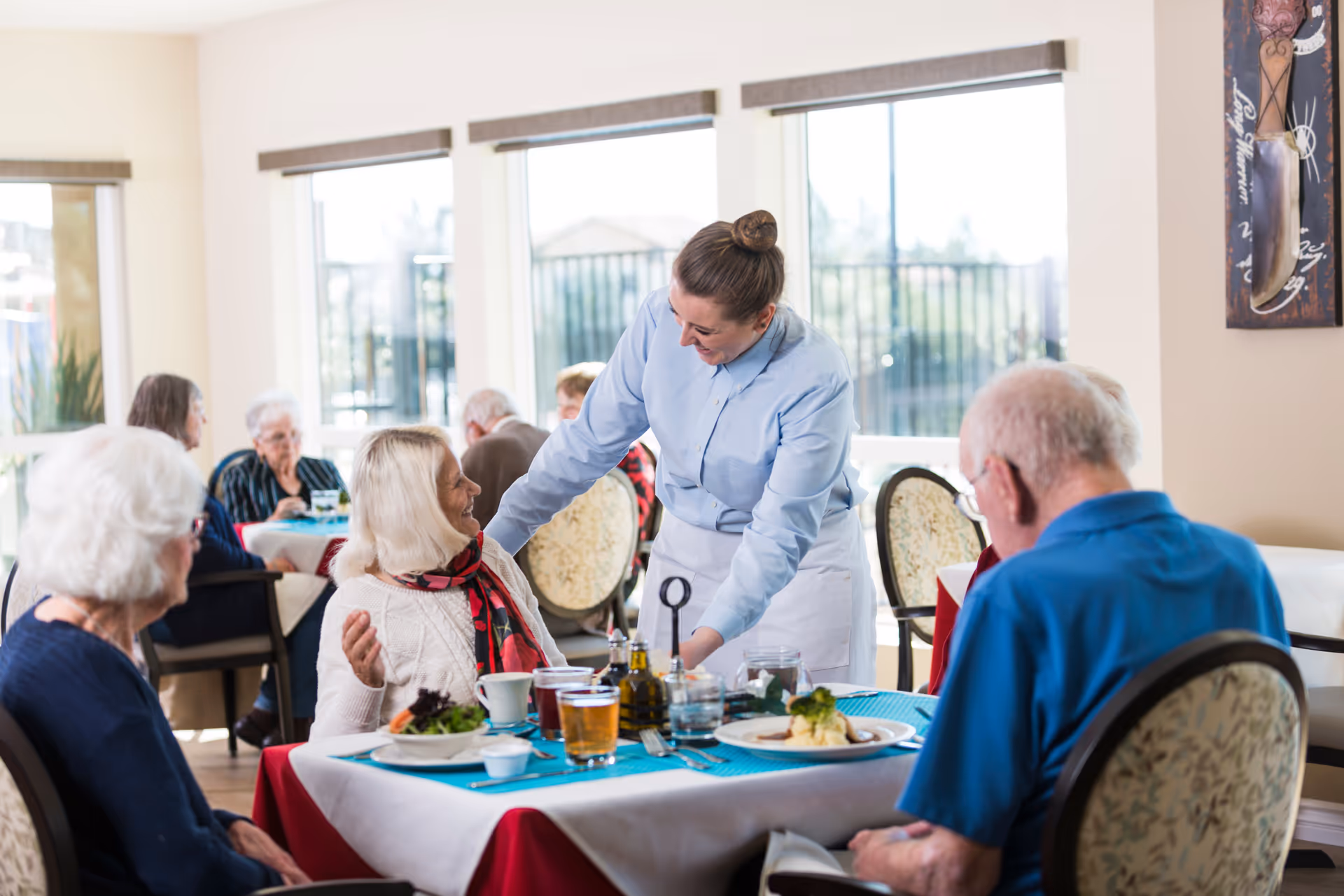 A dining room in a senior living facility where elderly residents are seated at tables enjoying their meals. A smiling waitress in a light blue shirt and white apron is interacting warmly with a seated elderly woman wearing a white sweater and red scarf. The room is bright with large windows letting in natural light.