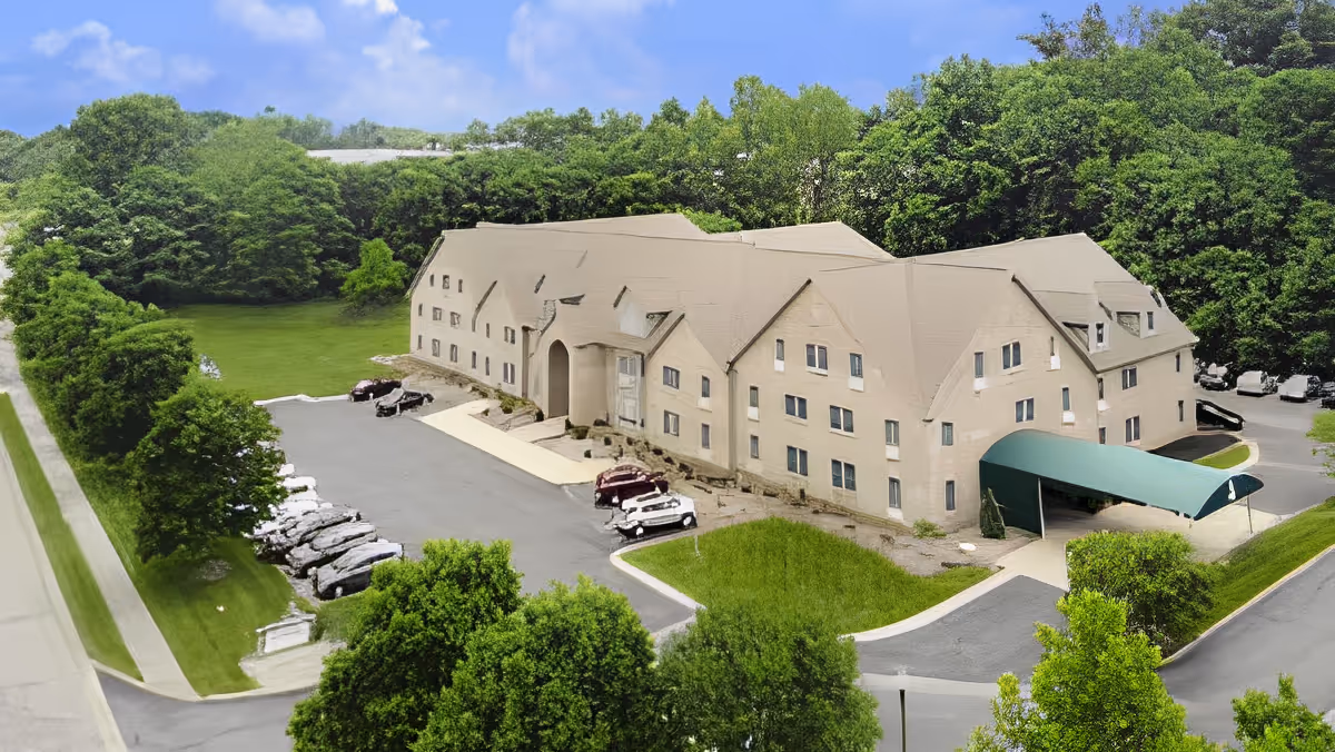 Aerial view of a large beige multi-story building with a green canopy over the entrance, surrounded by trees and greenery. There is a parking lot with several cars parked, and a road running alongside the property under a blue sky with some clouds.