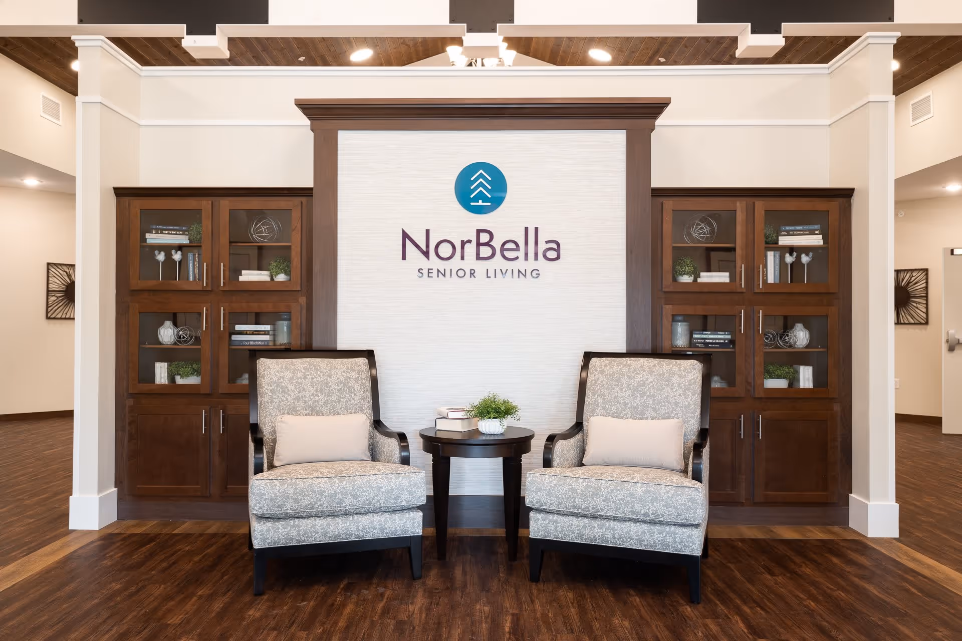 Two upholstered armchairs with light patterned fabric and beige cushions are positioned on either side of a small round wooden table with a plant and books on it. Behind the chairs is a wall with a NorBella Senior Living logo, flanked by wooden cabinets with glass doors containing decorative items and books. The floor is wooden, and the ceiling has recessed lighting.