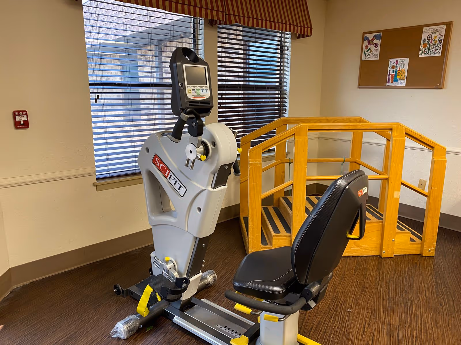 Rehabilitation room with a SciFit exercise machine, a recumbent bike seat, and wooden practice stairs beside a window.
