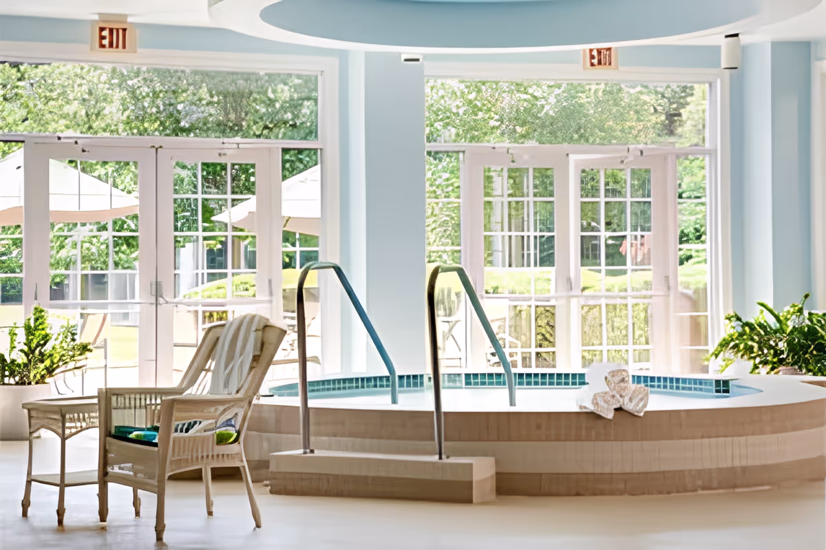 Indoor spa area with a round hot tub featuring metal handrails and steps. There are two wicker chairs with cushions and a small wicker table nearby. Large glass doors and windows in the background reveal a green outdoor garden with patio umbrellas and chairs. The walls are painted light blue and there are potted plants on either side of the hot tub.
