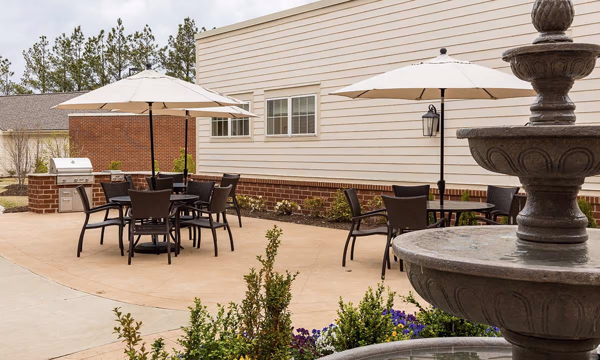 Outdoor patio area with round tables and chairs under large white umbrellas, a brick grill station, and a multi-tiered stone water fountain in the foreground. The patio is adjacent to a building with beige siding and windows, surrounded by some greenery and flowers.