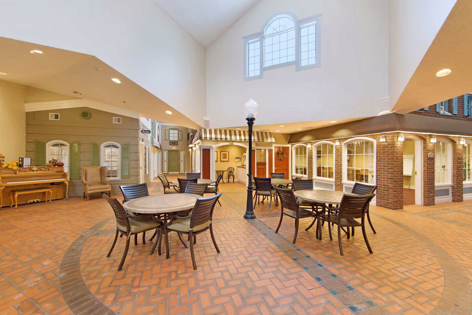 Indoor common area with brick flooring and several round tables with chairs arranged around a central lamp post. The space has high ceilings with a large window, and the walls are designed to look like the exterior of houses with windows, doors, and awnings. There is a piano and an armchair on the left side.