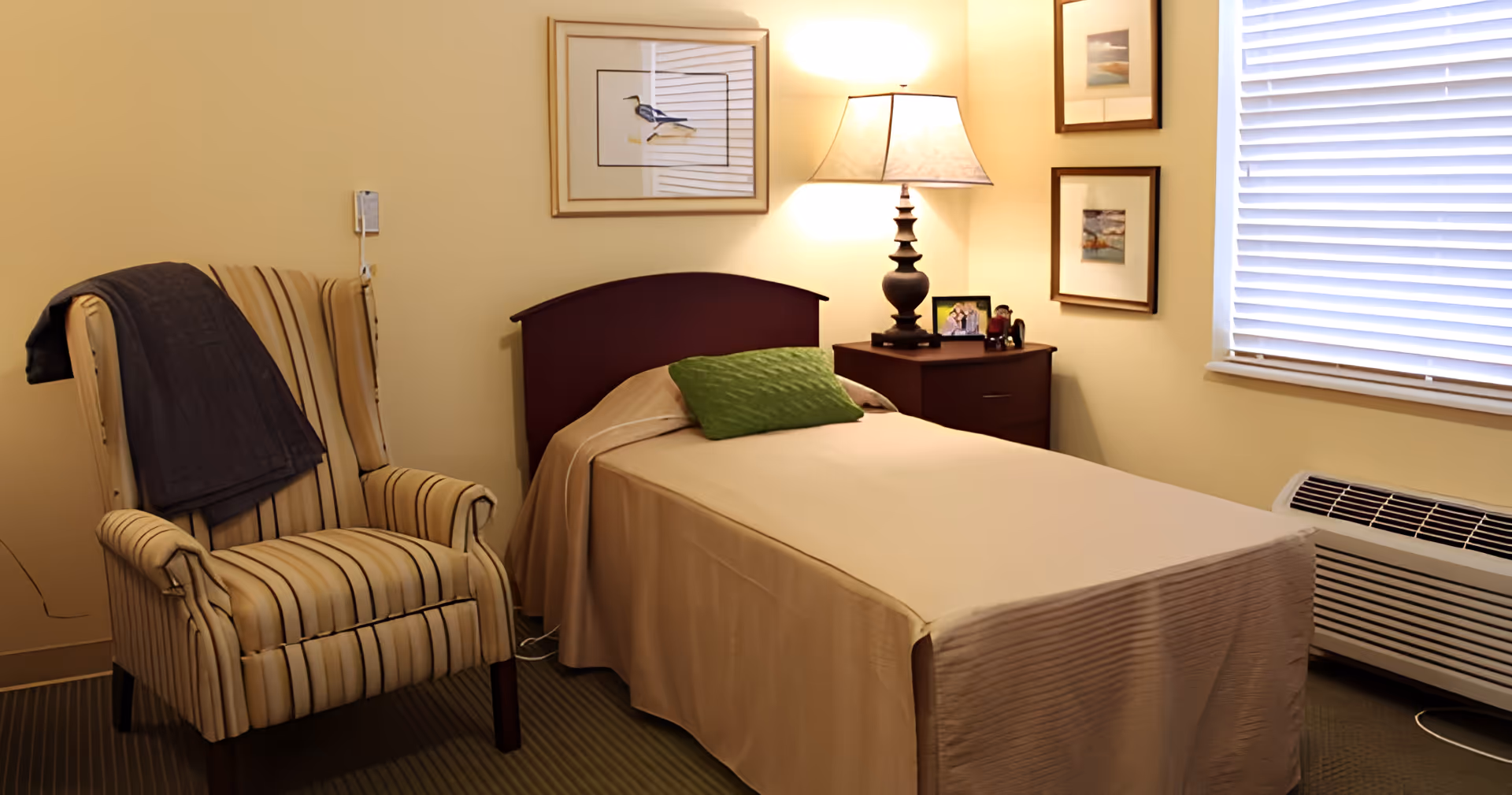 A tidy single-bed bedroom with a striped armchair, nightstand and lamp beside a window with blinds.