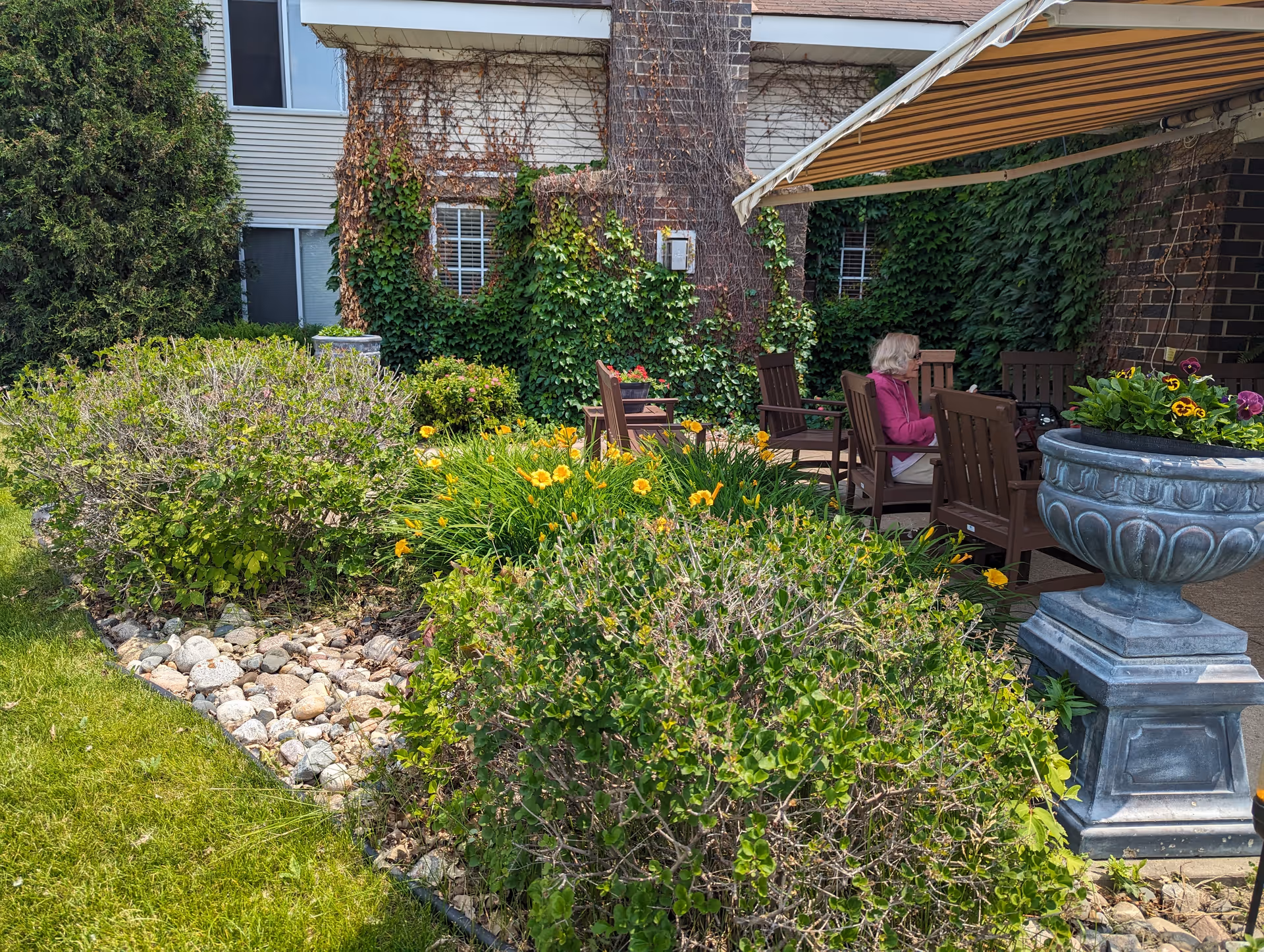 Outdoor patio area at a senior living facility with wooden chairs arranged around a table. A woman in a pink jacket is seated on one of the chairs. The patio is surrounded by green bushes, yellow flowers, and a large stone planter with colorful flowers. The building wall behind is covered with ivy and has windows.