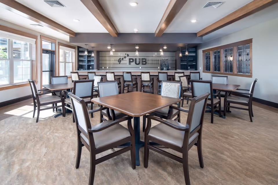 Interior view of a well-lit pub area with multiple wooden tables and chairs arranged neatly. The back wall features a bar counter with high chairs and shelves stocked with glasses. Large windows on the left side allow natural light to fill the room, and wooden beams run across the ceiling.