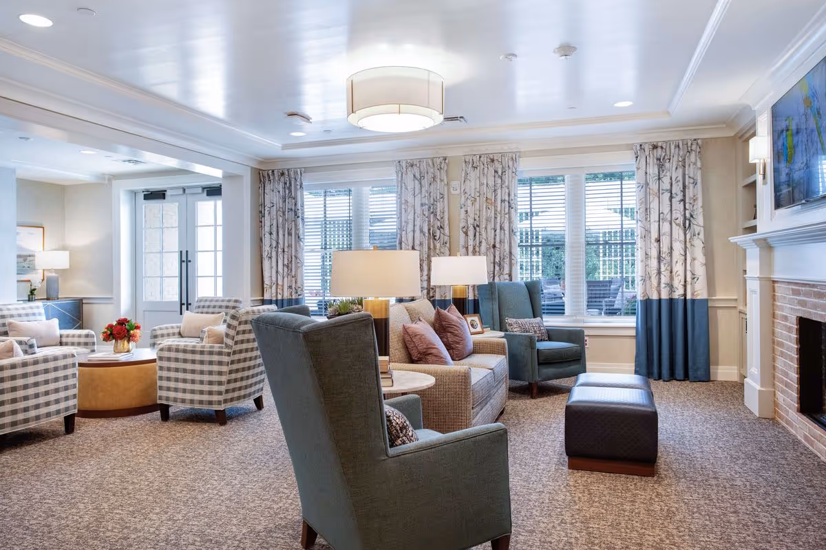 A bright and cozy living room area in a senior living facility with comfortable armchairs, a sofa with cushions, a round coffee table with a flower arrangement, large windows with patterned curtains, a fireplace, and a wall-mounted TV.