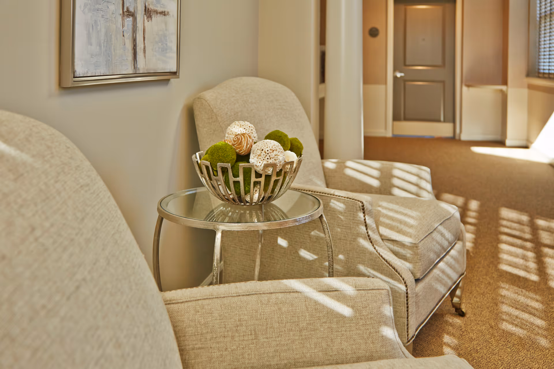 Two beige upholstered armchairs with a small round glass-top side table between them holding a decorative bowl with green and white ornamental balls. The setting is a well-lit hallway with sunlight streaming through window blinds, casting shadows on the carpeted floor and chairs. A closed door is visible at the end of the hallway.