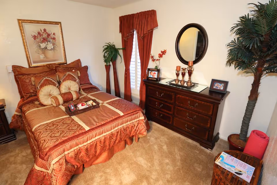 A cozy bedroom with a bed dressed in red and gold patterned bedding, including decorative pillows. Next to the bed is a wooden nightstand with a lamp. A wooden dresser with six drawers is topped with three candle holders, two framed photos, and a round mirror hanging above it. The room has beige carpet, red curtains on the window, a framed floral painting on the wall, and two tall potted plants in the corners. A wicker basket with magazines and a rolled-up pink yoga mat are also visible.