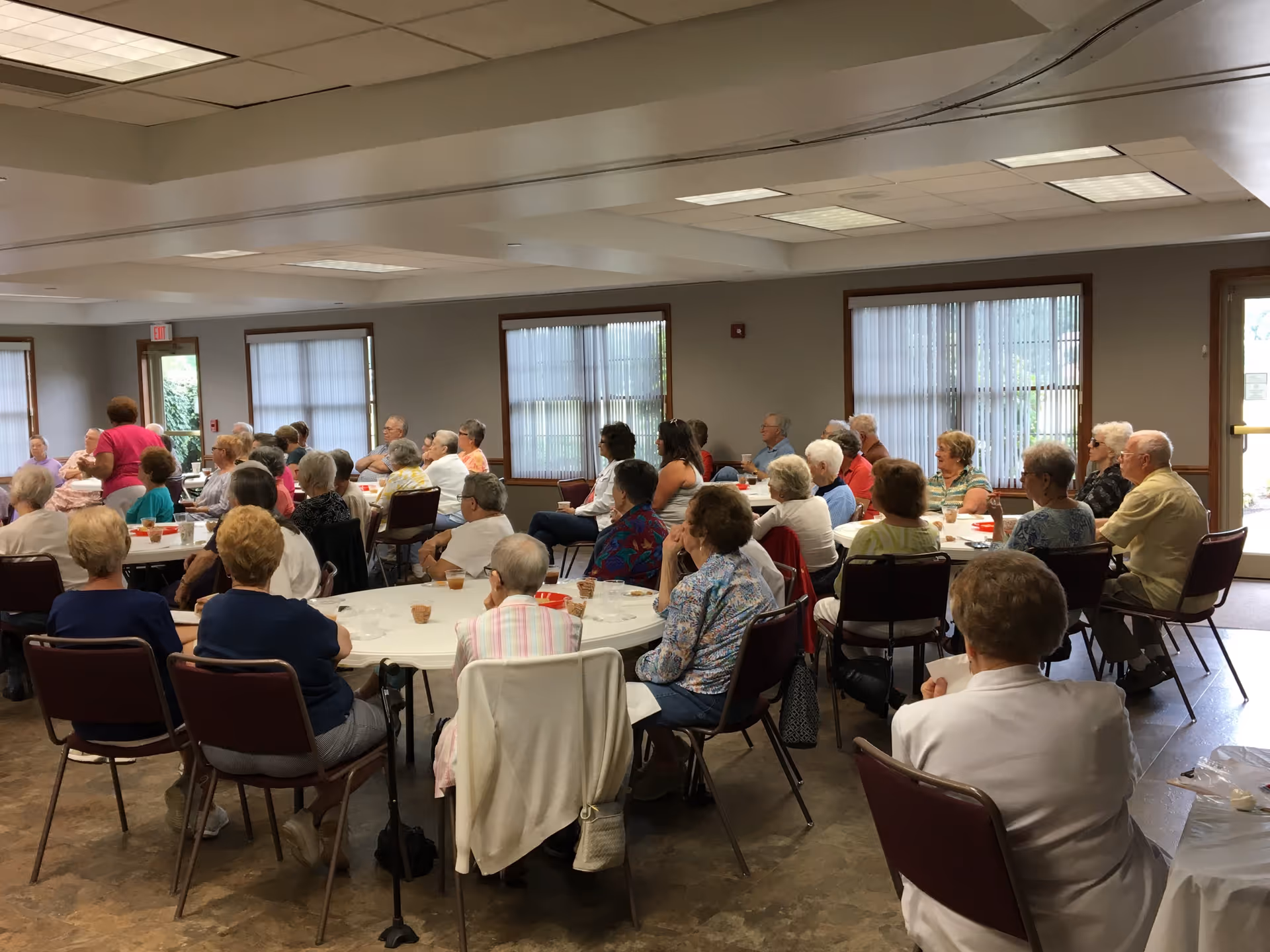 A group of elderly people seated around tables in a large room, attentively listening to a speaker. The room has windows with blinds, fluorescent ceiling lights, and a door leading outside.