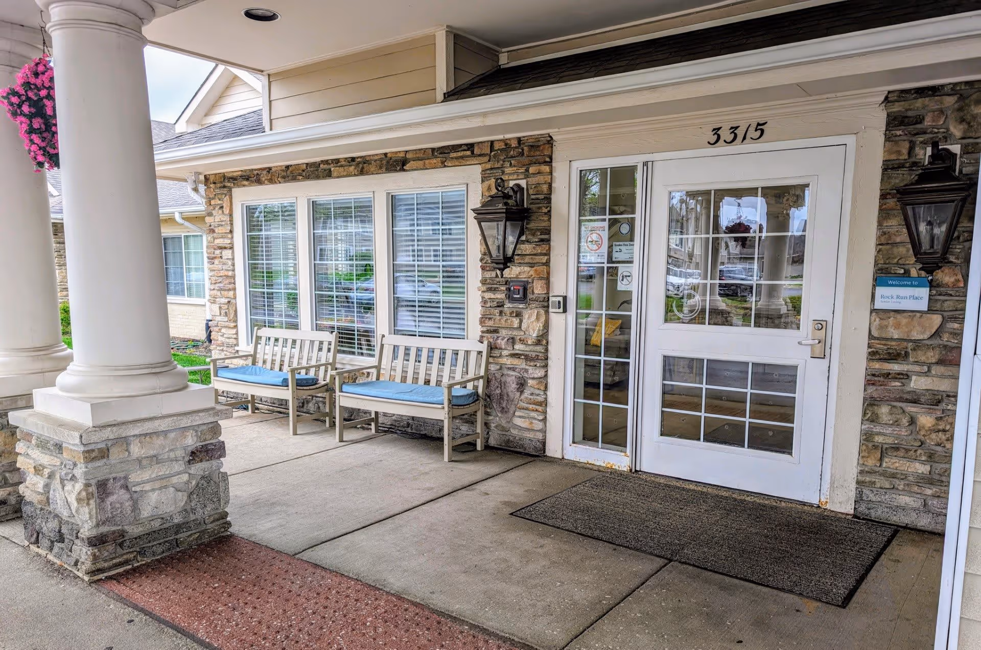 Covered front entrance of Rock Run Memory Care with stone pillars, two benches with blue cushions and a glass entry door marked 3315.
