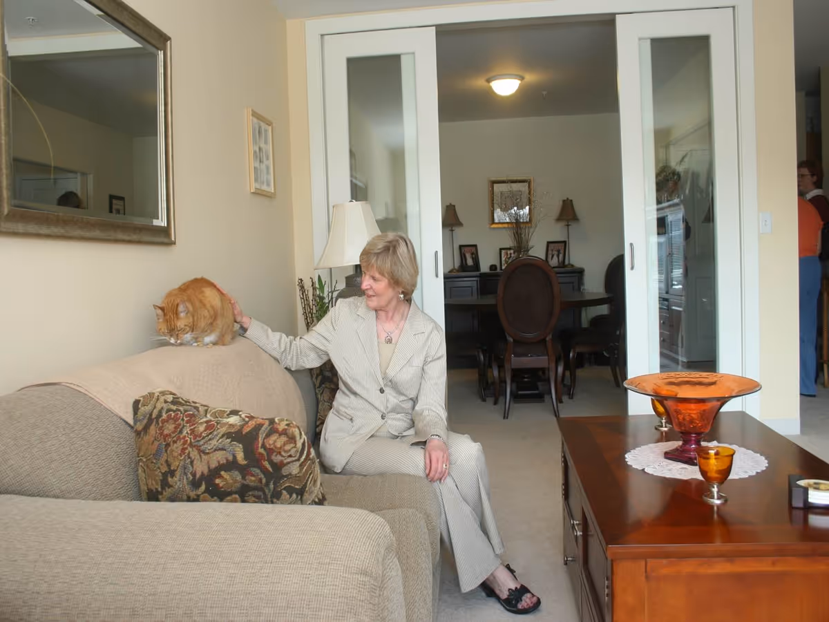 A woman in a light-colored suit sitting on a beige couch petting an orange cat that is perched on the back of the couch in a living room. The room has a large mirror on the wall, a wooden coffee table with decorative glass bowls, and sliding glass doors leading to a dining area with a round table and chairs.