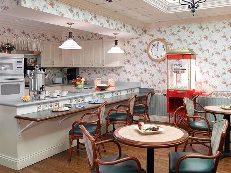 A cozy dining area in a senior living facility with a kitchen counter and bar stools. The kitchen has white cabinets, a microwave, and a coffee urn. There are round tables with chairs, a red popcorn machine, floral wallpaper, and a wall clock showing 12:05.
