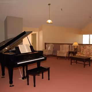 A spacious room with a black grand piano and a piano bench in the foreground. Behind the piano, there are two upholstered armchairs, a floral-patterned sofa, a wooden coffee table, and a side table with a lamp. The room has beige walls, a pink carpet, and a hanging ceiling light.