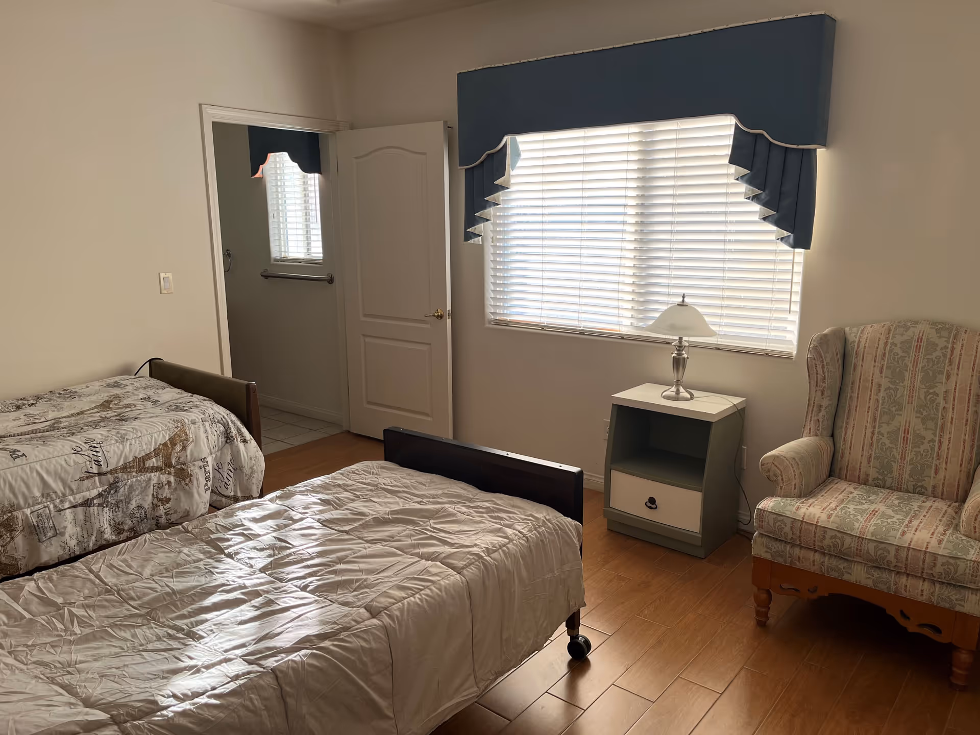 A simple bedroom with two single beds, one with a beige comforter and the other with a patterned cover featuring the Eiffel Tower. There is a window with closed blinds and a blue valance above it. Next to the window is a small nightstand with a lamp, and to the right is an upholstered armchair with a floral pattern. An open door reveals a glimpse of a bathroom with tiled flooring and a towel bar.