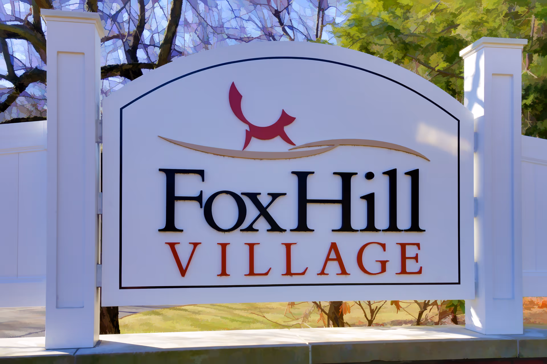 A white sign with black and red text reading 'Fox Hill Village' mounted between two white pillars with trees and grass in the background.