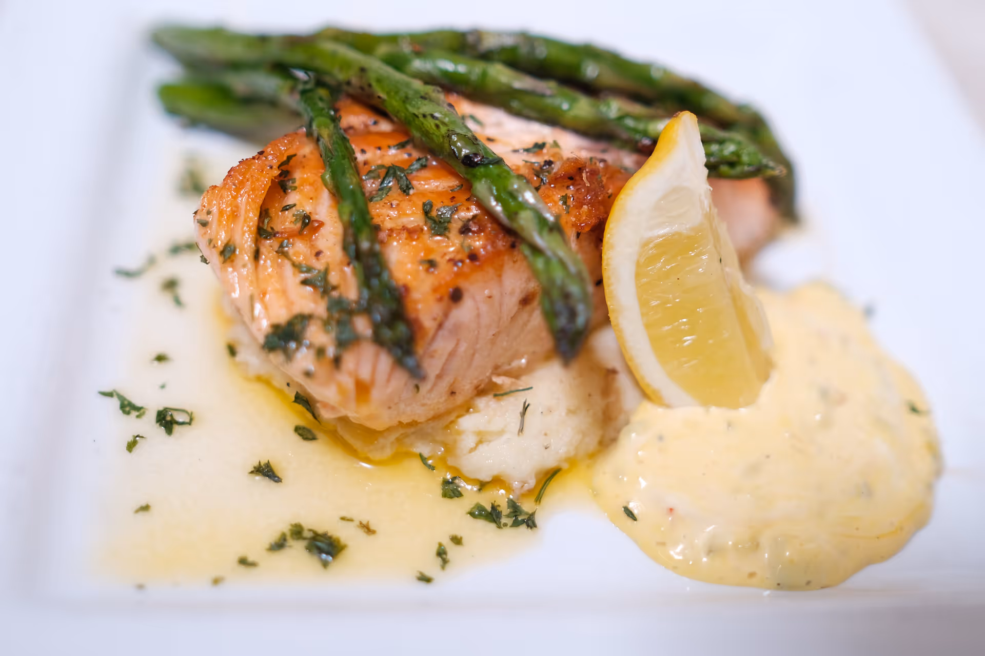 A plated meal featuring a cooked salmon fillet topped with grilled asparagus, served on a bed of mashed potatoes with a lemon wedge and a side of creamy sauce.