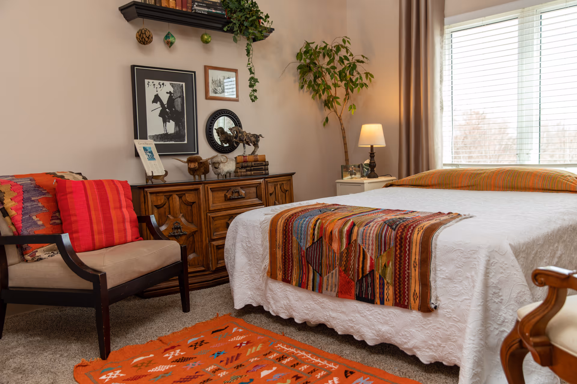 Cozy bedroom with a bed covered in a white quilt and colorful throw, a wooden dresser, an armchair with red cushions, and a window with blinds.