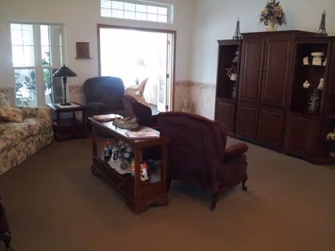A living room with a floral patterned sofa, two armchairs, a wooden coffee table with decorative items, a wooden entertainment cabinet with shelves holding various ornaments, and a windowed door leading to a bright adjacent room.