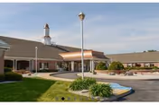 Exterior view of Carlisle Skilled Nursing and Rehabilitation Center showing a single-story building with a covered entrance, a circular driveway, landscaped greenery, and a clear sky.