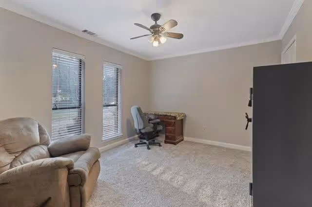 A small room with beige walls and carpeted floor featuring a comfortable recliner chair, a wooden desk with a swivel office chair, two tall windows with blinds, a ceiling fan with lights, and a large black safe against one wall.