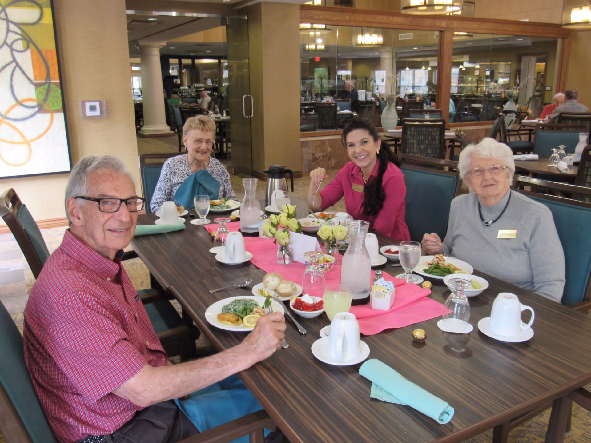 Four people sitting around a dining table in a retirement facility, enjoying a meal together. The table is set with plates of food, glasses of water, coffee cups, and a pink table runner with small flower arrangements. The background shows a spacious dining area with other tables and chairs.