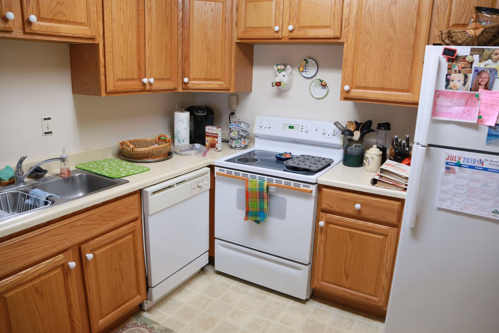 A compact kitchen with wooden cabinets, a white electric stove with a colorful towel hanging on the handle, a white dishwasher, and a white refrigerator covered with photos and notes. The countertop holds a paper towel roll, a coffee maker, a muffin mix box, a spice rack, kitchen utensils in a container, and some stacked plates. The floor has a light-colored linoleum pattern.