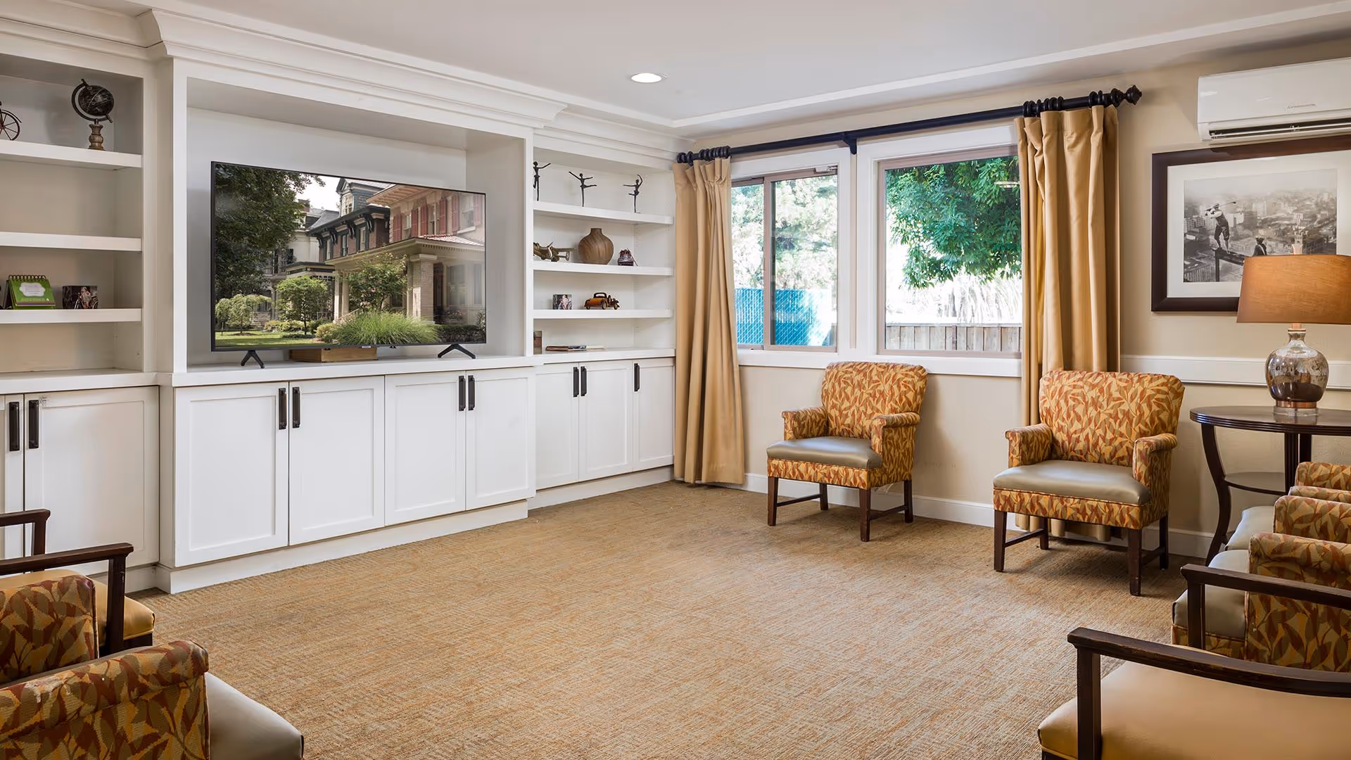 A bright and spacious living room with beige carpet, white built-in cabinets and shelves, a flat-screen TV, two windows with beige curtains, and several patterned armchairs arranged around the room. A table with a lamp and a framed black and white photo are also visible.