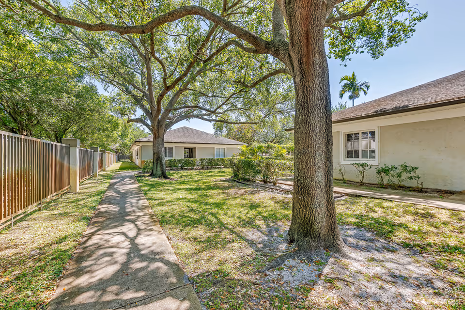 Sidewalk through a shaded grassy courtyard between single-story buildings with large oak trees.