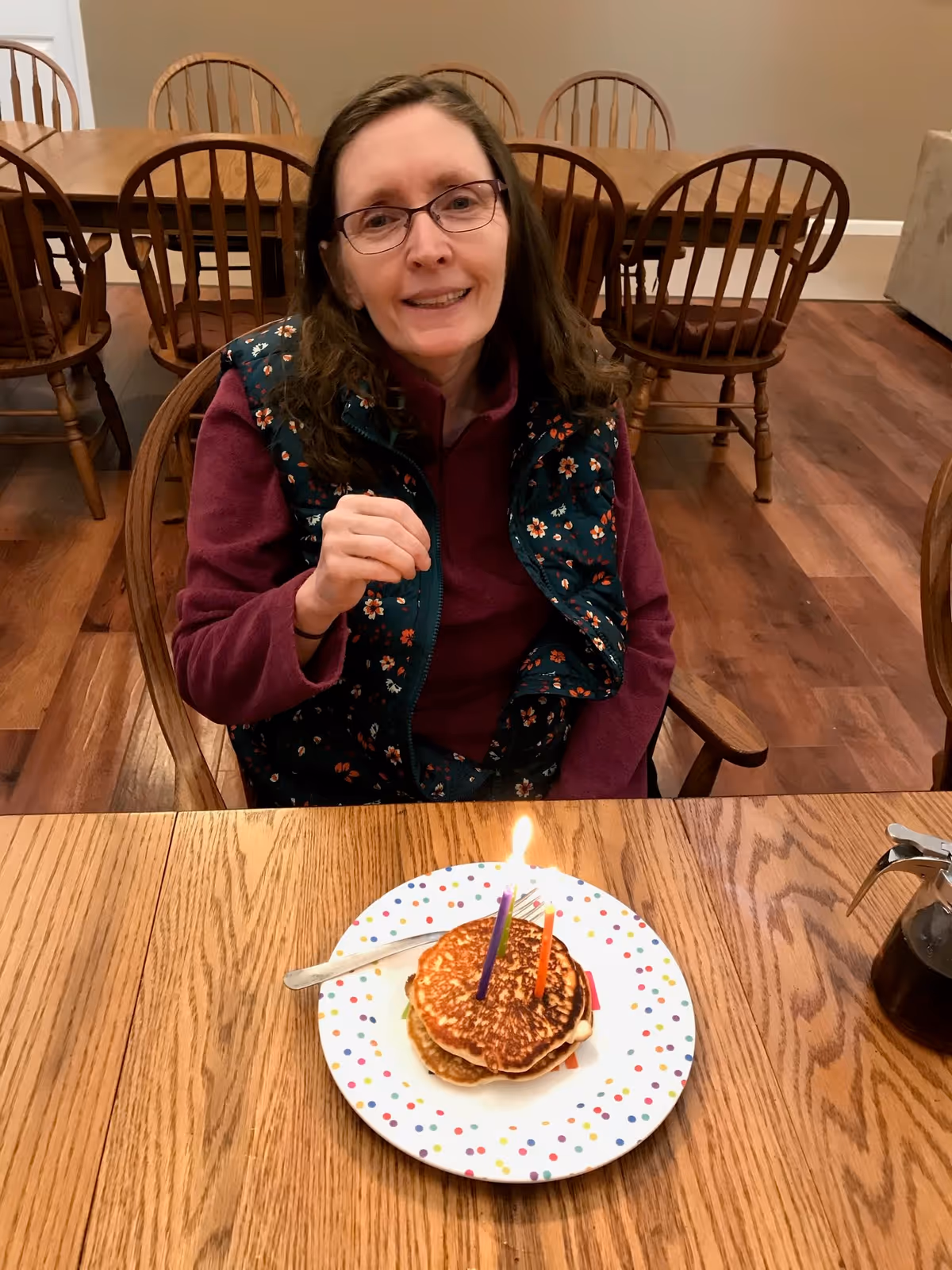 A woman with glasses and a floral vest sitting at a wooden table with a plate of pancakes topped with two lit birthday candles in front of her. The background shows wooden chairs and tables in a dining area.