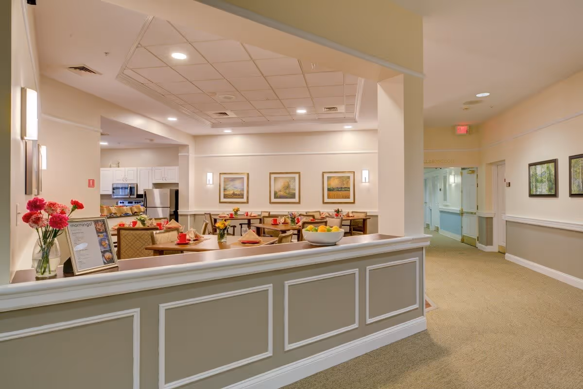 Interior view of a dining area in a senior living facility with tables set with red cups, napkins, and small flower arrangements. The room has beige walls, framed landscape paintings, and recessed lighting. A kitchen area with white cabinets and stainless steel appliances is visible in the background. A hallway with additional framed pictures and an exit sign is seen to the right.