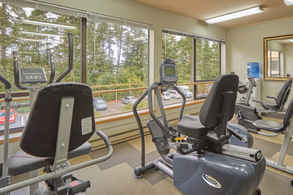 Exercise room with recumbent stationary bikes and cardio machines facing large windows overlooking trees and a parking lot.