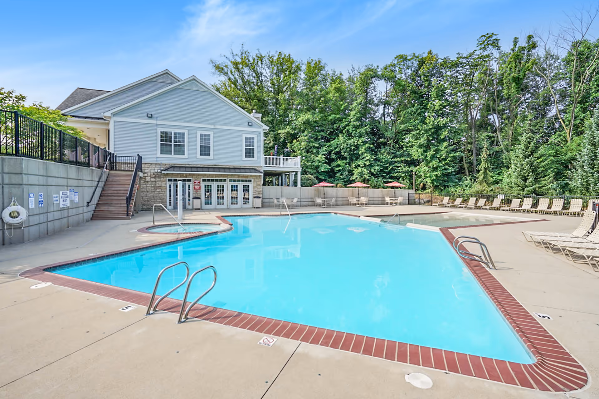 Outdoor swimming pool area with clear blue water, surrounded by a concrete deck with lounge chairs and tables with red umbrellas. A two-story building with light blue siding and stone accents is visible in the background, along with green trees and a clear blue sky.