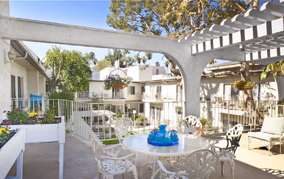 Outdoor courtyard patio with white wrought-iron table and chairs under a pergola overlooking apartment balconies and hanging plants.