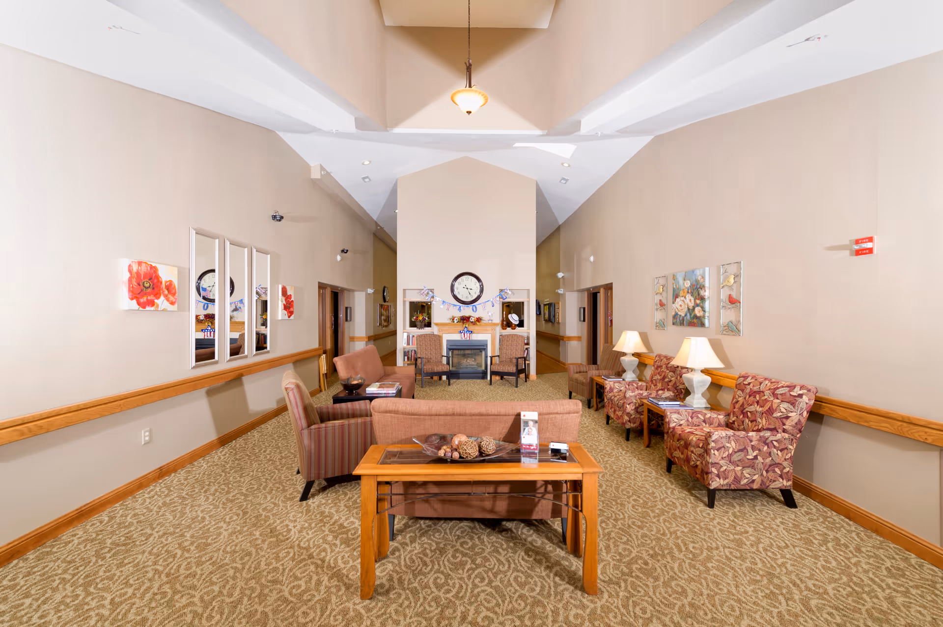 A spacious and well-lit common area with beige walls and carpeted floor. The room features multiple seating options including two patterned armchairs with lamps on side tables, a striped armchair, and two sofas facing a fireplace with a clock above it. Decorative paintings and mirrors adorn the walls, and a wooden table with decorative items is placed in the foreground.