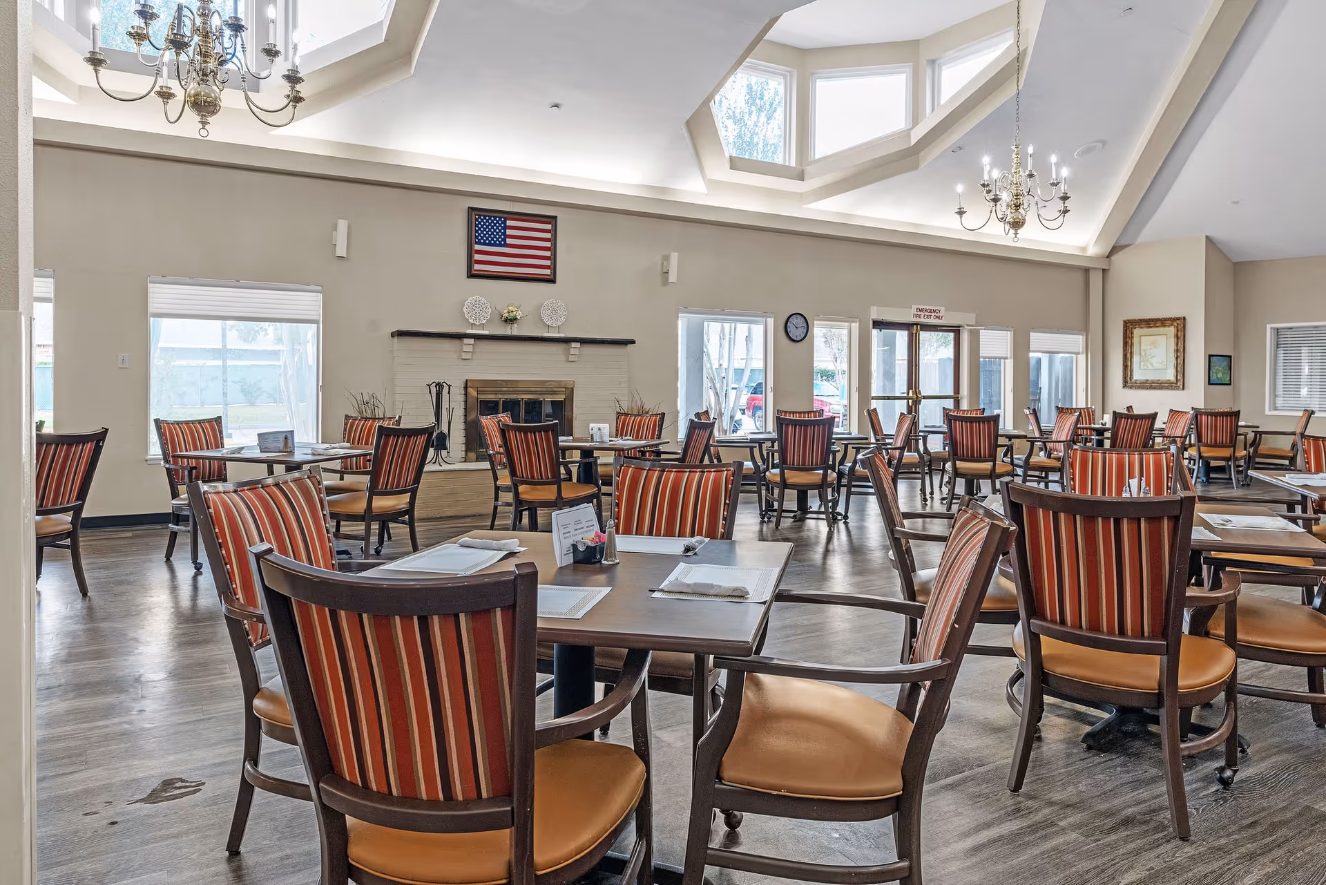 Spacious dining room with multiple tables and red-striped chairs, chandeliers, skylight, and a fireplace.