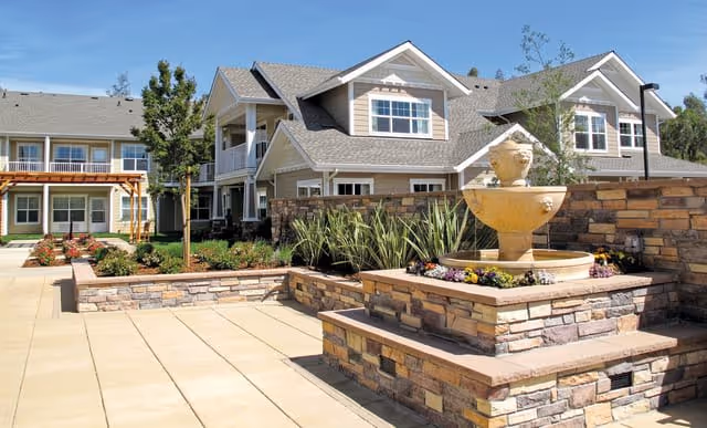 Outdoor courtyard area of a senior living facility with a stone fountain centerpiece surrounded by flower beds and plants. The background shows a two-story building with beige siding, white trim, and multiple windows under a clear blue sky.