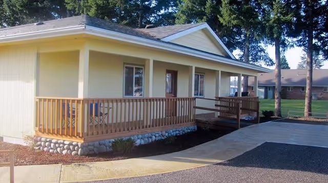 Exterior view of a single-story yellow building with a wooden porch railing and a small ramp leading to the entrance, surrounded by trees and a paved driveway.