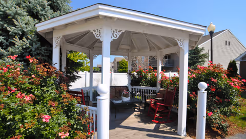 A white wooden gazebo surrounded by blooming rose bushes and greenery under a clear blue sky. Inside the gazebo, there are red rocking chairs and a small table with a basket on it. A lamppost and residential buildings are visible in the background.