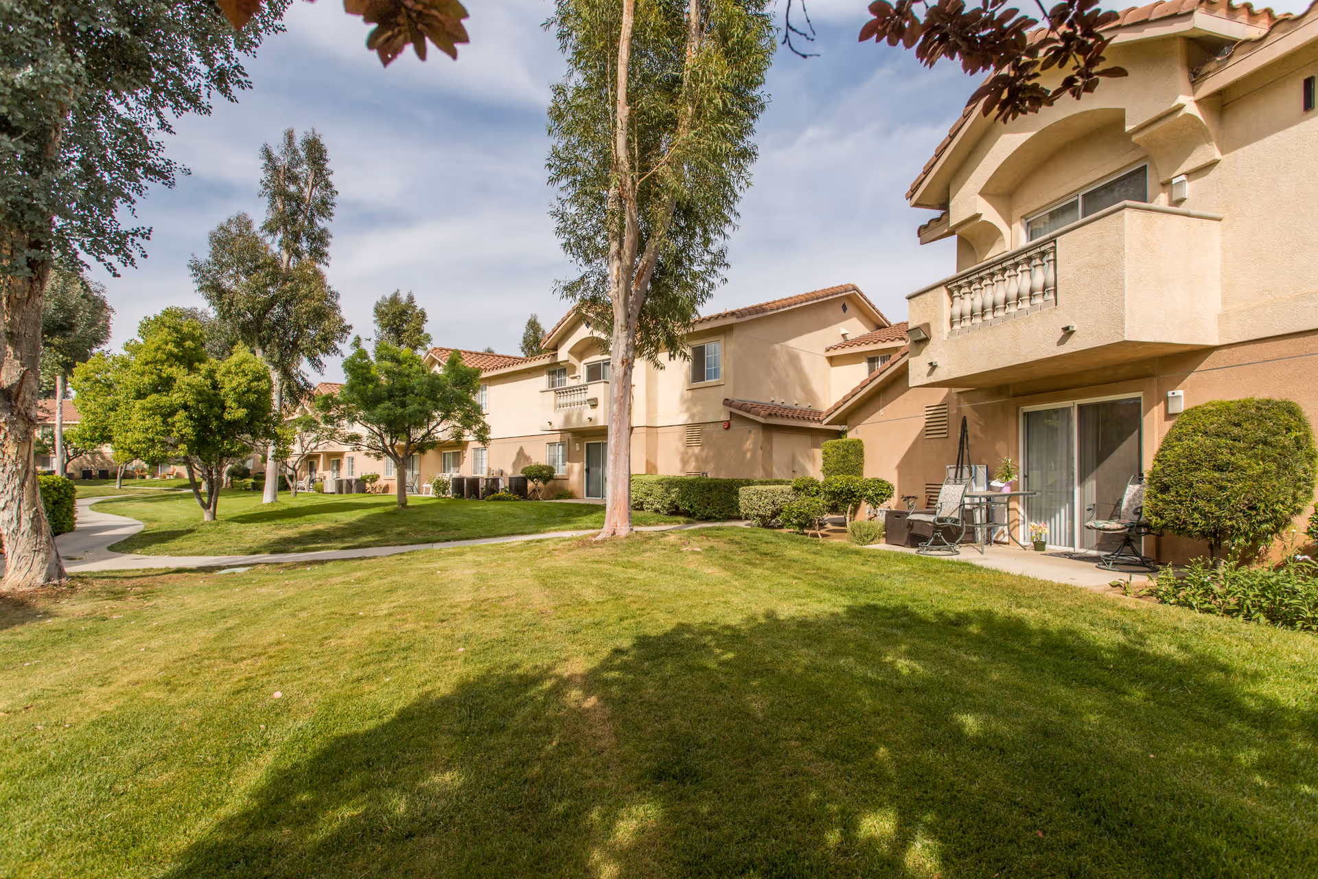 Outdoor view of a senior living facility with beige stucco buildings featuring balconies and patios. The area has well-maintained green lawns, trees, and shrubs, with a paved walkway winding through the landscape under a partly cloudy sky.