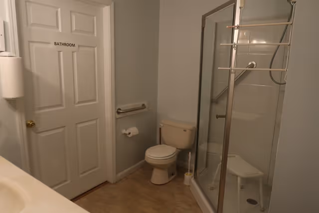 Small bathroom with a labeled door, toilet, glass-enclosed shower with a shower chair and grab bars.