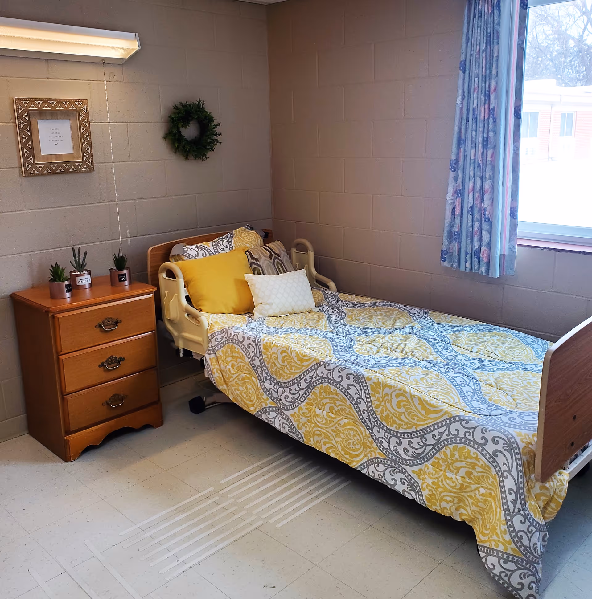 A neatly made single bed with yellow patterned bedding beside a wooden nightstand and window in a simple senior living bedroom.