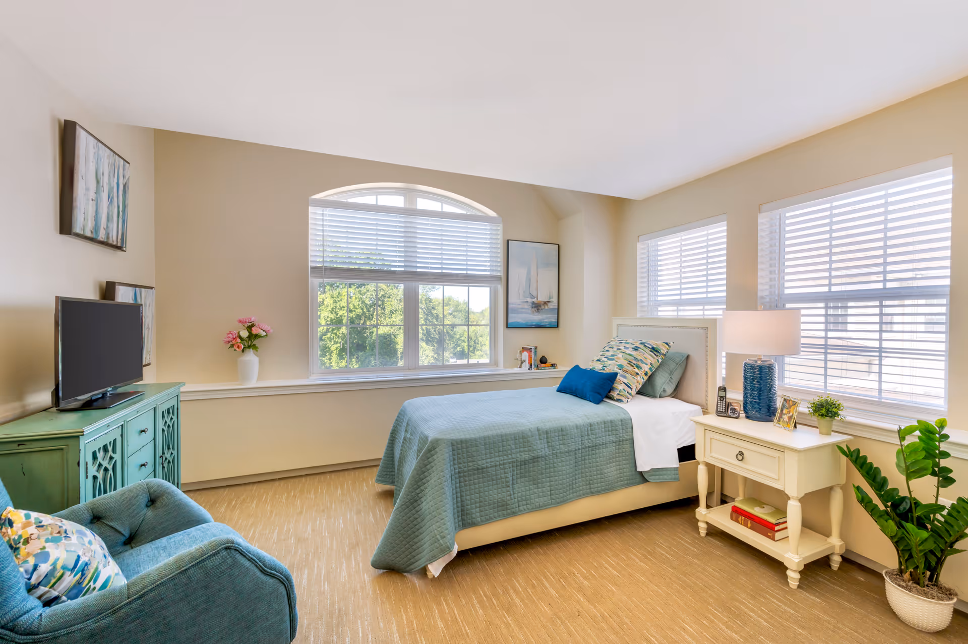 A bright and cozy bedroom with a single bed covered in a teal quilt and decorative pillows. The room features large windows with white blinds allowing natural light to fill the space. There is a white nightstand with a blue lamp, a small plant, a phone, and framed photos. A teal armchair with a colorful cushion and a teal cabinet with a TV on top are also present. The walls are painted beige and decorated with framed artwork. A potted plant is placed on the floor near the nightstand.